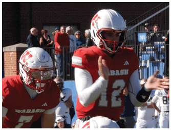 A group of people in red and white football uniforms are lined up on a green field preparing for a play with a large brick building and spectators in the background. Description generated by AI