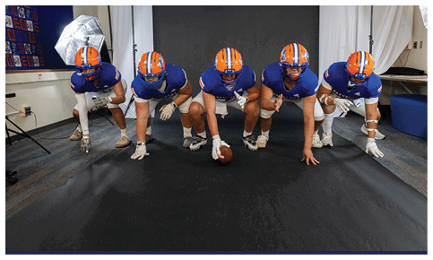 Several people wearing athletic shoes and gloves are positioned on a black surface with one person holding a football in a ready stance. Description generated by AI