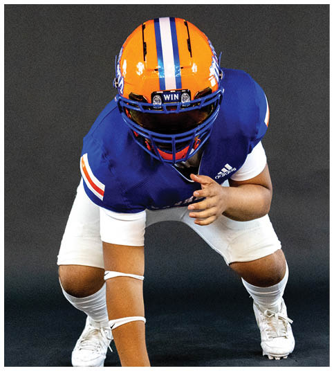 A person wearing a blue and orange football uniform and helmet is in a crouched position against a dark background. Description generated by AI