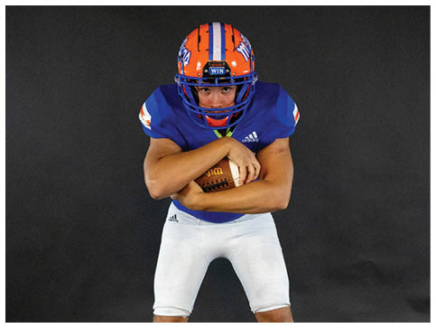 A person wearing a blue shirt and white pants is holding a football tightly against their body. Description generated by AI