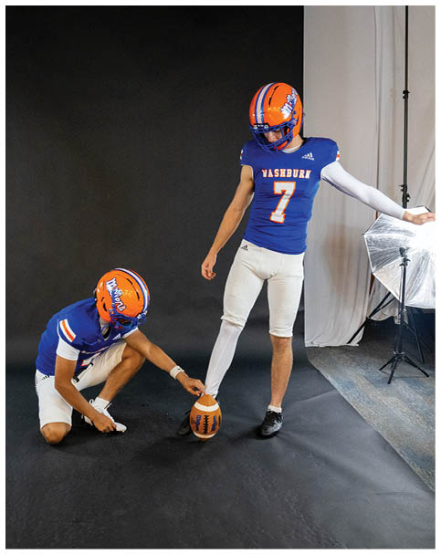 Two people wearing blue and orange football uniforms are posing in a studio with one person holding a football on the ground while the other stands with one foot on the ball. Description generated by AI