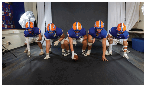 Several people wearing athletic shoes and gloves are positioned on a black surface with one person holding a football in a ready stance. Description generated by AI