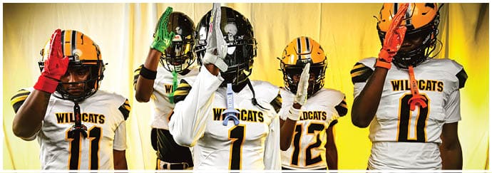 Five people wearing Wildcats football uniforms and helmets stand in a row with one hand raised in front of a yellow backdrop. Descripci n generada con IA