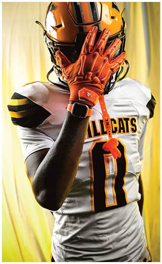 A person wearing an orange and black football helmet, orange gloves, and a white jersey is holding their hand up to their face while wearing a smartwatch. Descripci n generada con IA