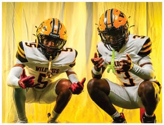 Two adults wearing yellow and white football uniforms with helmets are crouching in front of a yellow backdrop while making hand gestures. Descripci n generada con IA