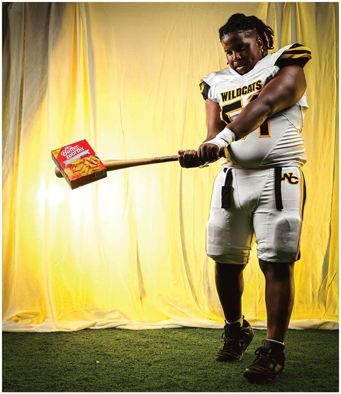 A person in a Wildcats football uniform is swinging a bat that has a box of Little Debbie Nutty Bars attached to the end against a yellow backdrop. Descripci n generada con IA