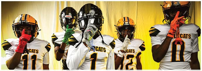 Five people wearing football helmets and uniforms stand in front of a yellow curtain, each holding a finger to their lips in a shushing gesture. Descripci n generada con IA