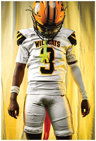 A person wearing a Wildcats football uniform with the number 3 on the jersey and a helmet is standing against a yellow background. Descripci n generada con IA