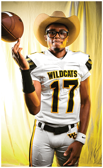 Person wearing a Wildcats football uniform with the number 17 and a cowboy hat is spinning a football on one finger against a yellow backdrop. Descripci n generada con IA