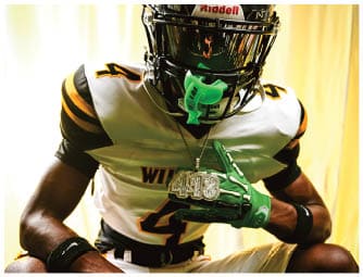 A person wearing a black football helmet, white and black football jersey with yellow stripes, green gloves, and a large chain necklace is posing against a light-colored background. Descripci n generada con IA