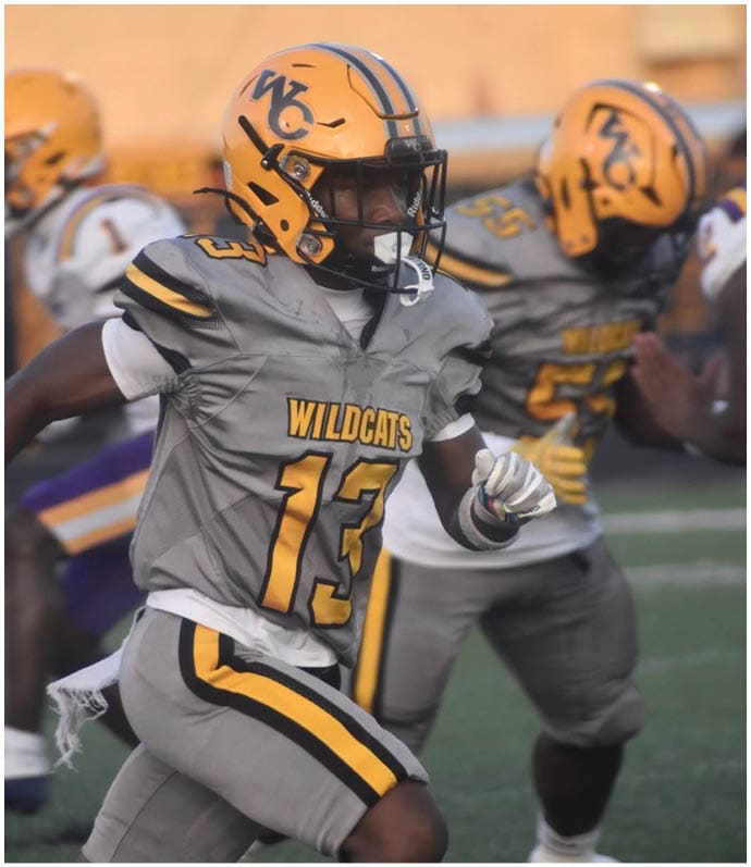 A person wearing a Wildcats football uniform with the number 13 is running on a field with other players in the background. Descripci n generada con IA