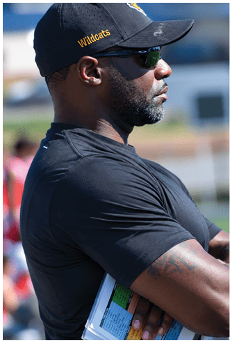 A person wearing a black Wildcats cap, sunglasses, and a black shirt is standing outdoors with arms crossed, looking to the side. Descripci n generada con IA