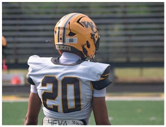 A person wearing a yellow football helmet with black and white stripes and the letters WC on the side is standing in front of empty bleachers. Descripci n generada con IA