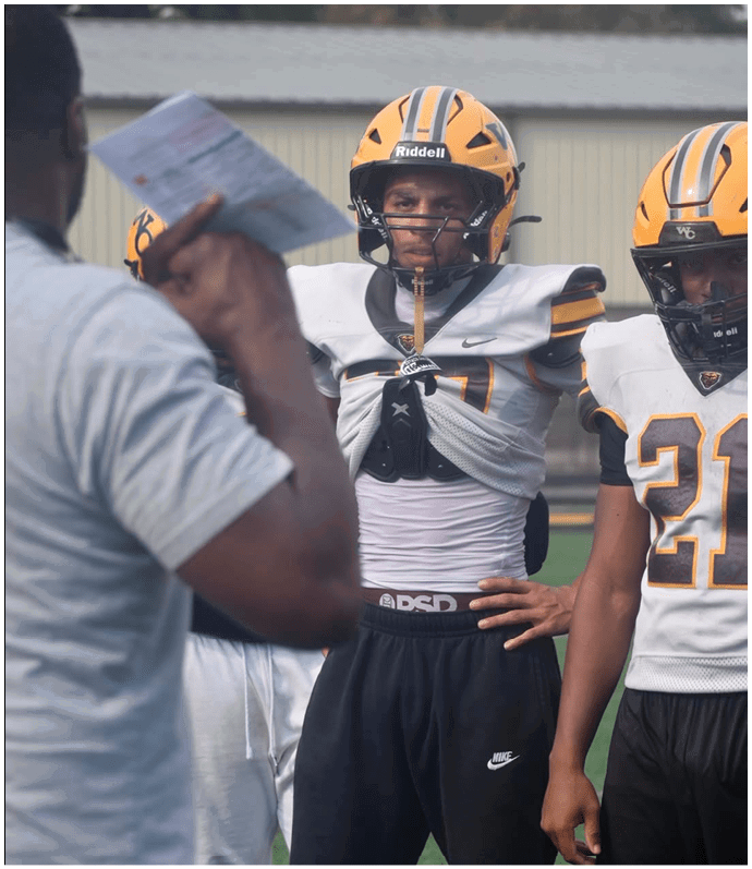 A person wearing a yellow and black football helmet and uniform stands with one hand on their hip while another person in a gray shirt holds a paper in the foreground. Descripci n generada con IA
