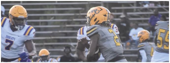 Two people in football uniforms are actively engaged on a field with bleachers and other players in the background. Descripci n generada con IA