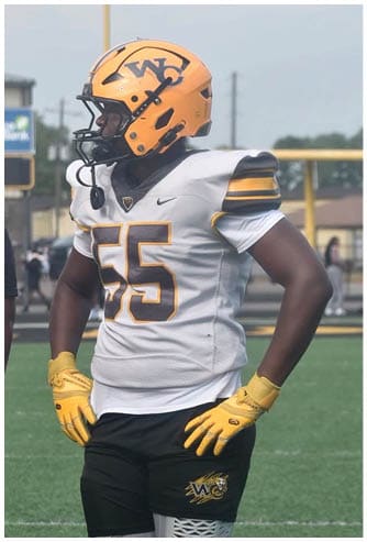 A person wearing a yellow football helmet, white jersey with the number 55, black shorts, and yellow gloves stands on a football field. Descripci n generada con IA