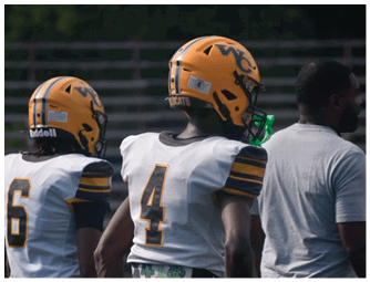 Two people wearing yellow football helmets and white jerseys with black and yellow stripes stand next to a person in a gray shirt on a sports field. Descripci n generada con IA