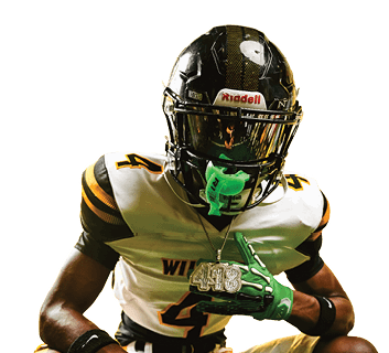 Person wearing a football uniform with a black helmet, green gloves, and neon green cleats is crouching on grass while holding a large chain necklace. Descripci n generada con IA