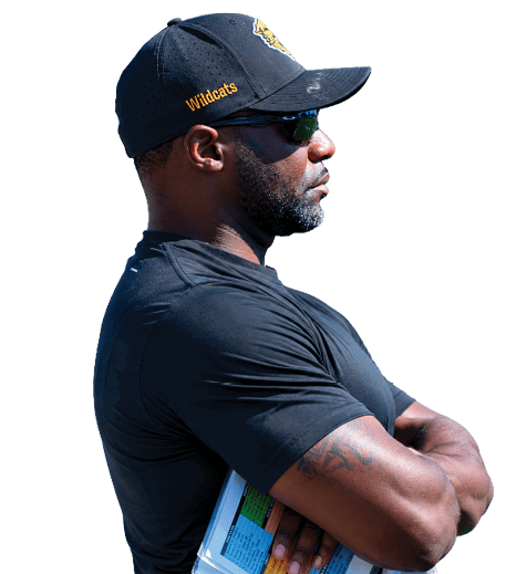 A person wearing a black Wildcats cap, sunglasses, and a black shirt stands with arms crossed holding a colorful clipboard outdoors. Descripci n generada con IA