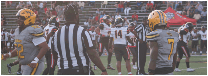 People in football uniforms and a referee are standing on a field during a game with spectators in the background. Descripci n generada con IA
