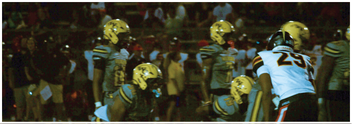 Several people wearing football helmets and uniforms are positioned on a field, appearing to prepare for a play during a nighttime game. Descripci n generada con IA