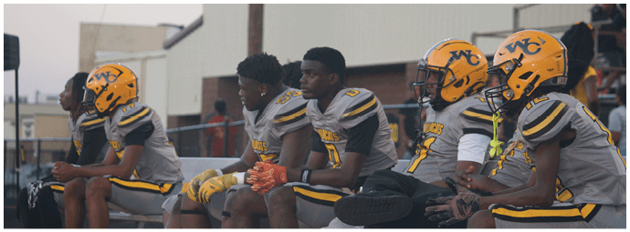 Several people wearing yellow and gray football uniforms and helmets are sitting on a bench on the sidelines of a sports field. Descripci n generada con IA