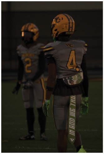 Two people wearing football uniforms and helmets stand on a field, with one facing away and the other facing forward in the background. Descripci n generada con IA
