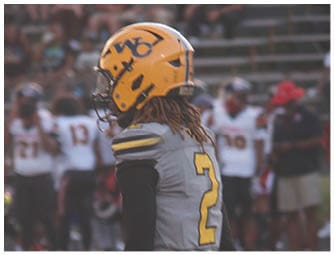 A person wearing a yellow helmet and gray football uniform with the number 2 stands on a football field with other players and coaches in the background. Descripci n generada con IA