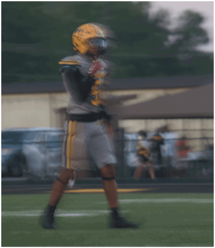 A person wearing a yellow helmet and gray sports uniform is standing on a field with a blurred background of trees and buildings. Descripci n generada con IA