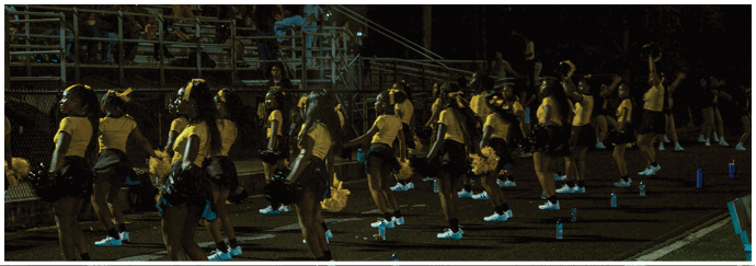 A group of people in matching outfits are performing a coordinated routine outdoors at night in front of a seated audience on bleachers. Descripci n generada con IA