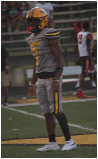 A person in a gray and yellow football uniform stands on a field while another person in a white and black uniform walks in the background. Descripci n generada con IA