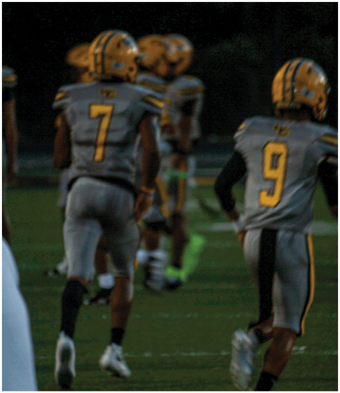 Two people in gray and yellow football uniforms with helmets are running on a field during a game. Descripci n generada con IA