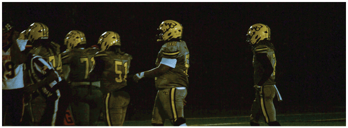 A group of football players wearing gold helmets and dark uniforms stand together on a field at night. Descripci n generada con IA