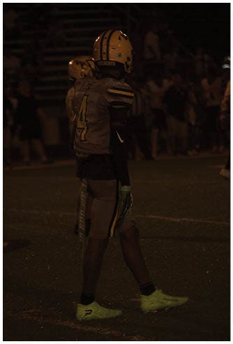 A person wearing a football uniform and helmet stands on a dimly lit field with other people blurred in the background. Descripci n generada con IA