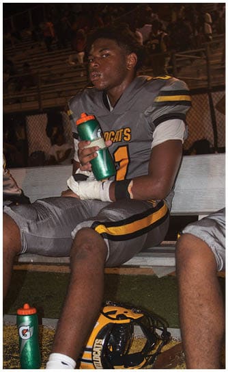 A person in a Wildcats football uniform is sitting on a bench holding a green water bottle with another water bottle and a helmet on the ground nearby. Descripci n generada con IA