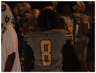 People wearing football helmets and uniforms are gathered closely together in a dimly lit setting. Descripci n generada con IA