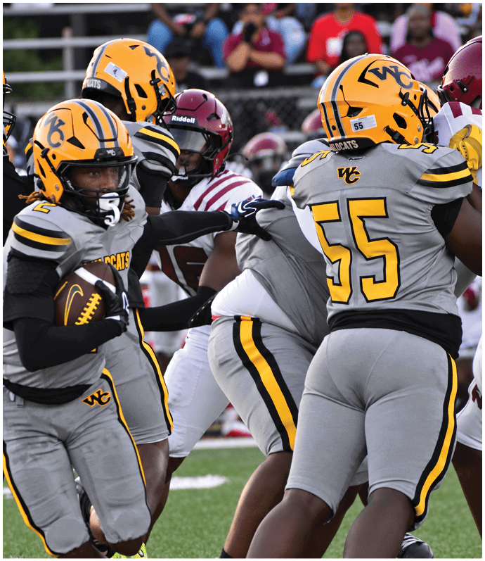 A person wearing a Wildcats football uniform is kicking a ball on a field with a blurred spectator in the background. Descripci n generada con IA