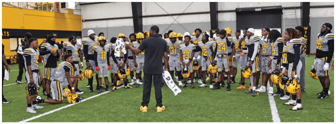 A group of people wearing yellow and gray football uniforms are gathered indoors around a person in a black shirt who appears to be addressing them. Descripci n generada con IA