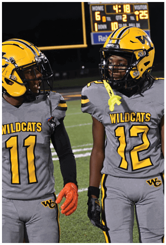 Two people wearing football helmets and uniforms are on a field with a scoreboard visible in the background. Descripci n generada con IA