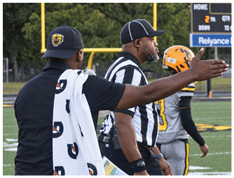 Two people wearing sports attire and hats are standing on a field near a goalpost, with one person holding a towel and the other wearing a striped referee shirt and headset. Descripci n generada con IA