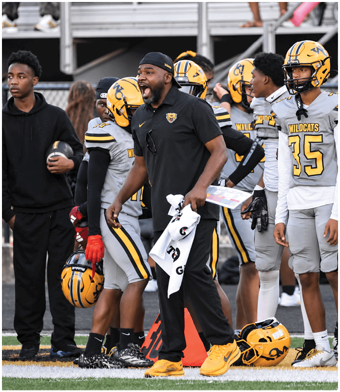 A group of people in football uniforms and a coach in black sportswear are standing on the sidelines of a football field, with the coach appearing to shout instructions. Descripci n generada con IA