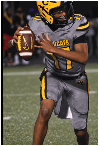 A person wearing a yellow and gray football uniform with a helmet is preparing to throw a football on a field during a game. Descripci n generada con IA
