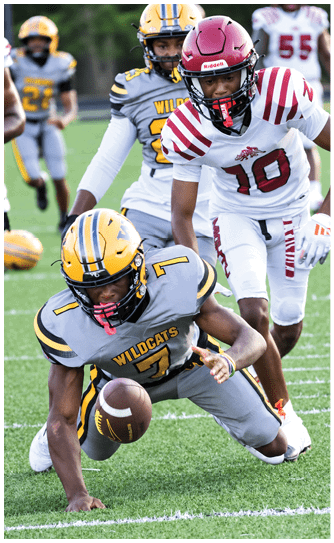 A person in a gray and yellow Wildcats football uniform is reaching down to catch a football on a grassy field while other players in different uniforms are nearby. Descripci n generada con IA