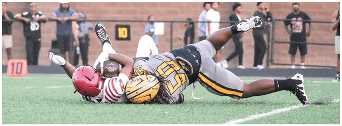 Two people in sports uniforms are lying on the ground during a football game with spectators standing behind a fence in the background. Descripci n generada con IA