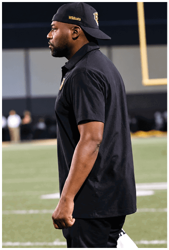 A person wearing a black shirt, black pants, and yellow Nike sneakers is walking on a football field near a goalpost. Descripci n generada con IA