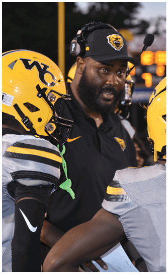 A person wearing a headset and cap with a tiger logo is talking to a football player in a yellow helmet and black and gray uniform. Descripci n generada con IA
