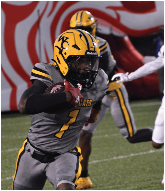A person in a gray and yellow football uniform is running with the ball on a field while two other players in different uniforms chase behind. Descripci n generada con IA