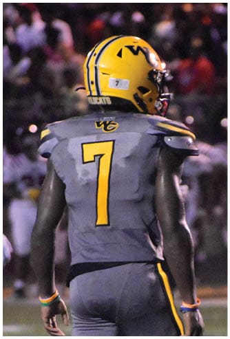 A person wearing a yellow helmet and gray football uniform with the number seven stands on a football field during a nighttime game. Descripci n generada con IA