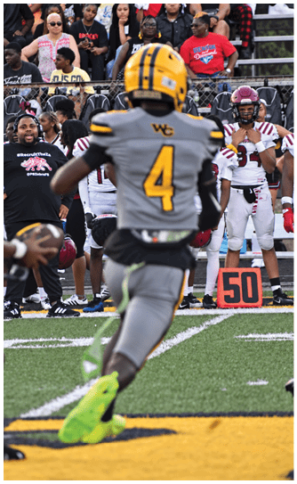 A person wearing a yellow helmet and gray football uniform with the number 4 is running on a football field toward a group of people and players standing near the sideline with a 50-yard marker visible. Descripci n generada con IA