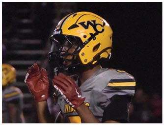 A person wearing a yellow football helmet and gray uniform with the number 5 is clapping on a football field at night with other players in the background. Descripci n generada con IA